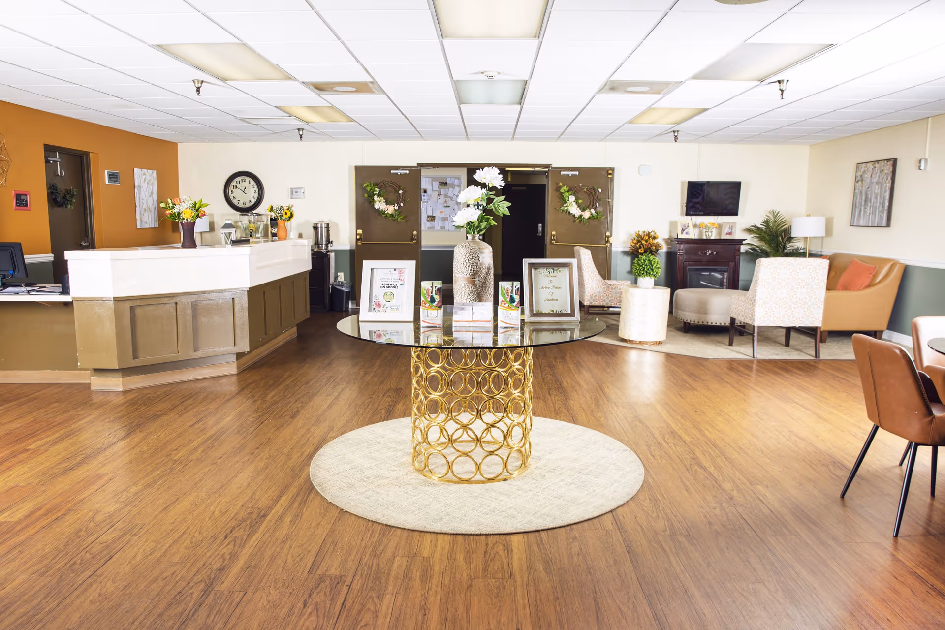 Bright and spacious senior living facility lobby with a round glass table featuring a gold circular base and a vase of white flowers in the center. To the left is a reception desk with flowers and a clock on the wall behind it. In the background, double doors decorated with wreaths lead to another room. To the right, there is a seating area with armchairs, a small table, a TV mounted on the wall, and a fireplace. The floor is wood with a light-colored round rug under the table.