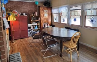 A cozy dining area with a round wooden table surrounded by four chairs, including two black metal chairs and two wooden chairs. The room has wooden flooring, a brick accent wall, a red cabinet, a wooden bookshelf, and windows decorated with hanging snowflake ornaments letting in natural light.
