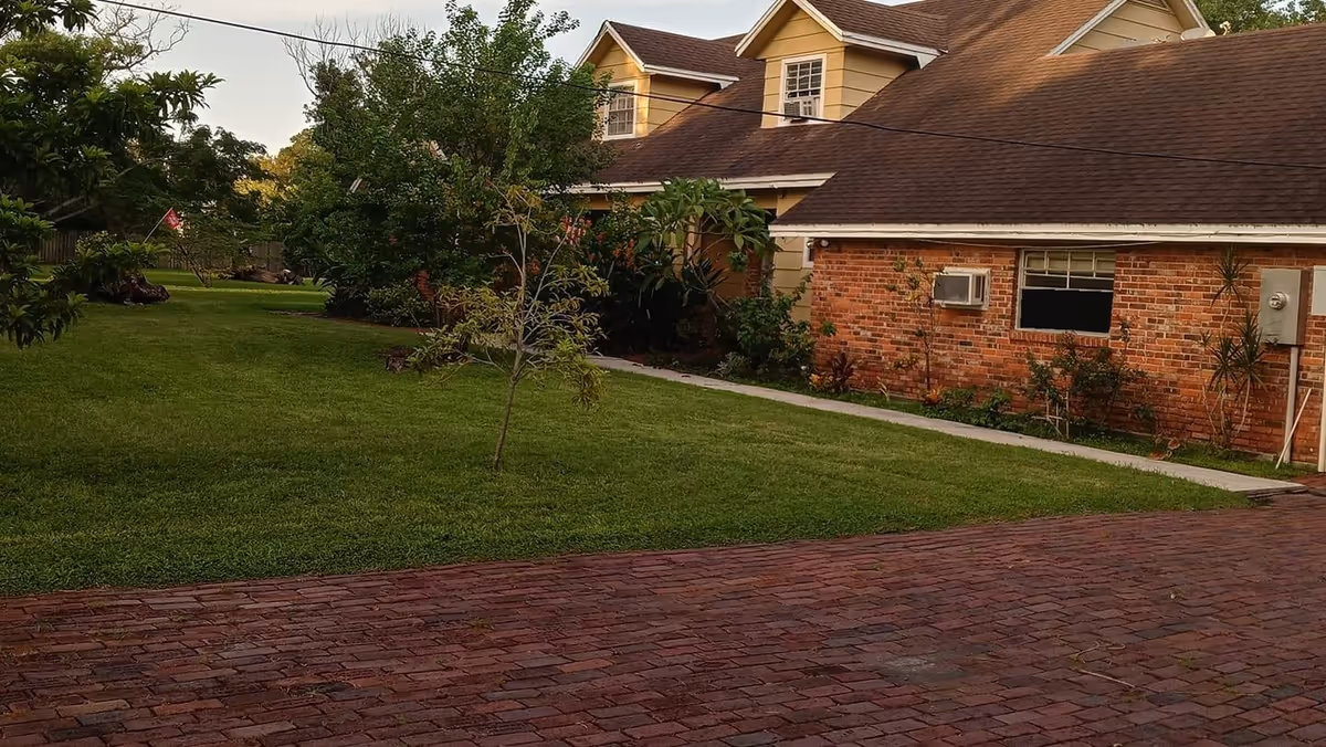 A brick-paved driveway leading to a brick and siding building with a brown shingled roof. The building is surrounded by a well-maintained green lawn with small trees and shrubs. The sky is clear and the scene is lit by natural daylight.