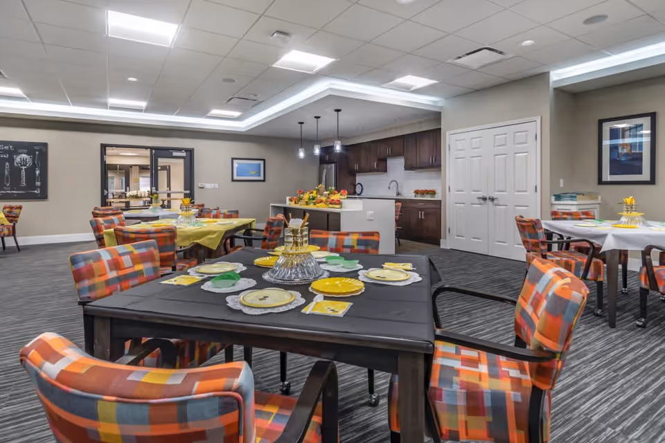 A senior living community dining area with multiple tables set for a meal. The tables have colorful checkered chairs and are covered with tablecloths and plates. In the background, there is a kitchen area with dark wood cabinets and a countertop decorated with fruit. The room has bright overhead lighting and neutral-colored walls with framed artwork.