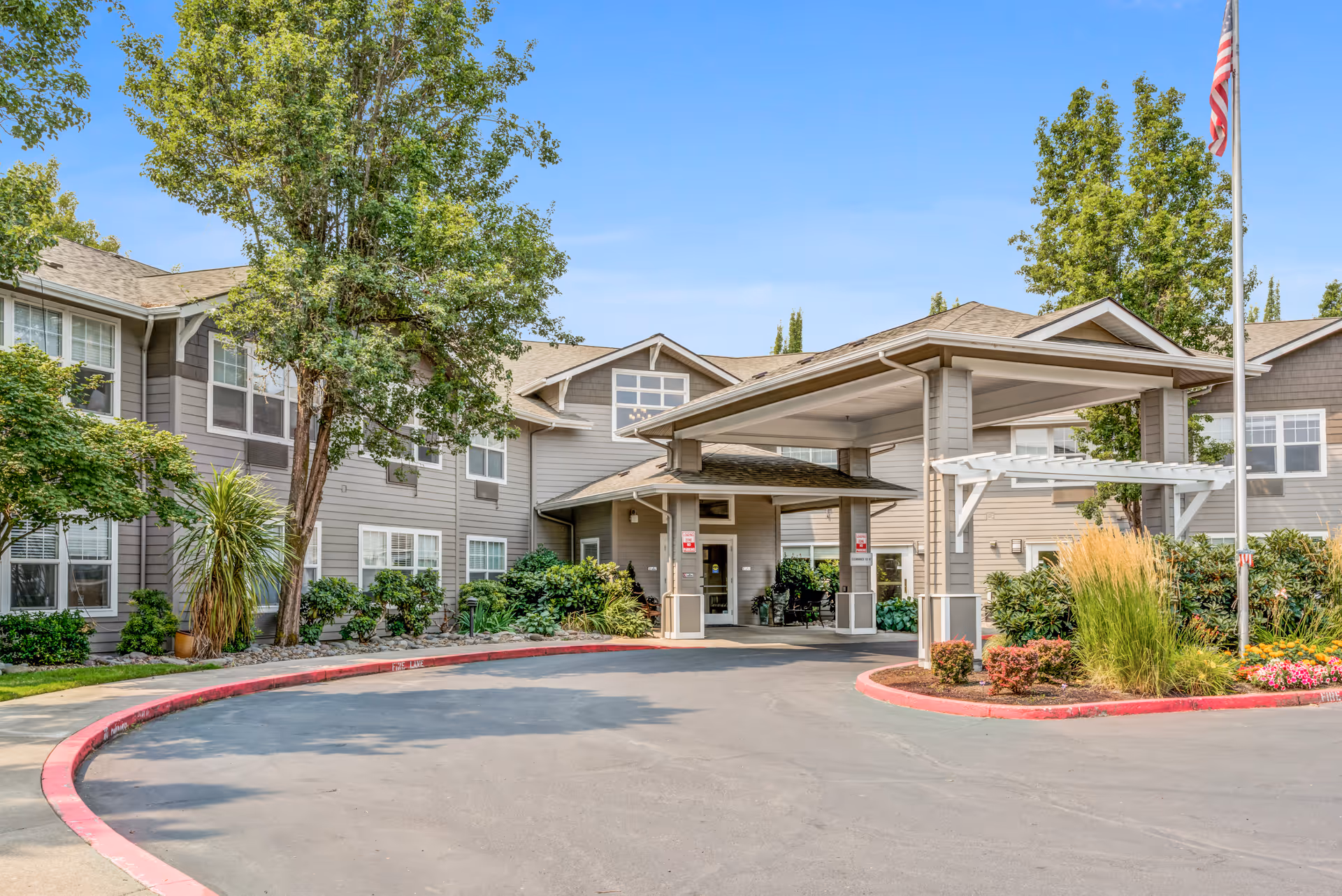 Exterior view of Woodside Senior Living facility showing a covered entrance with a driveway, surrounded by well-maintained landscaping including trees, shrubs, and flowers. The building is two stories with multiple windows and an American flag on a flagpole to the right.