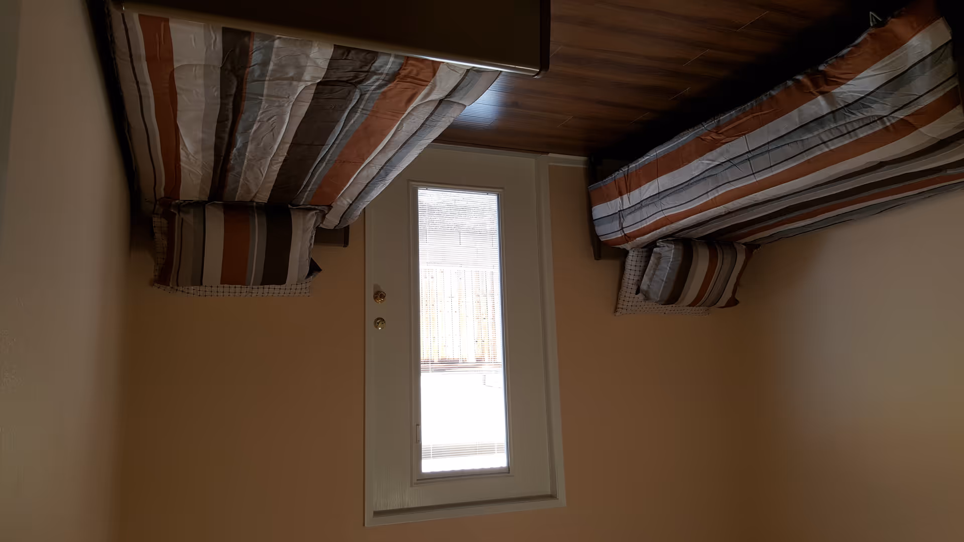 A small bedroom with two single beds placed parallel to each other against opposite walls. Each bed has striped bedding in shades of brown, gray, and white, with matching pillows. There is a door with a window in the center of the far wall, allowing natural light into the room. The floor is wooden, and the walls are painted a light beige color.