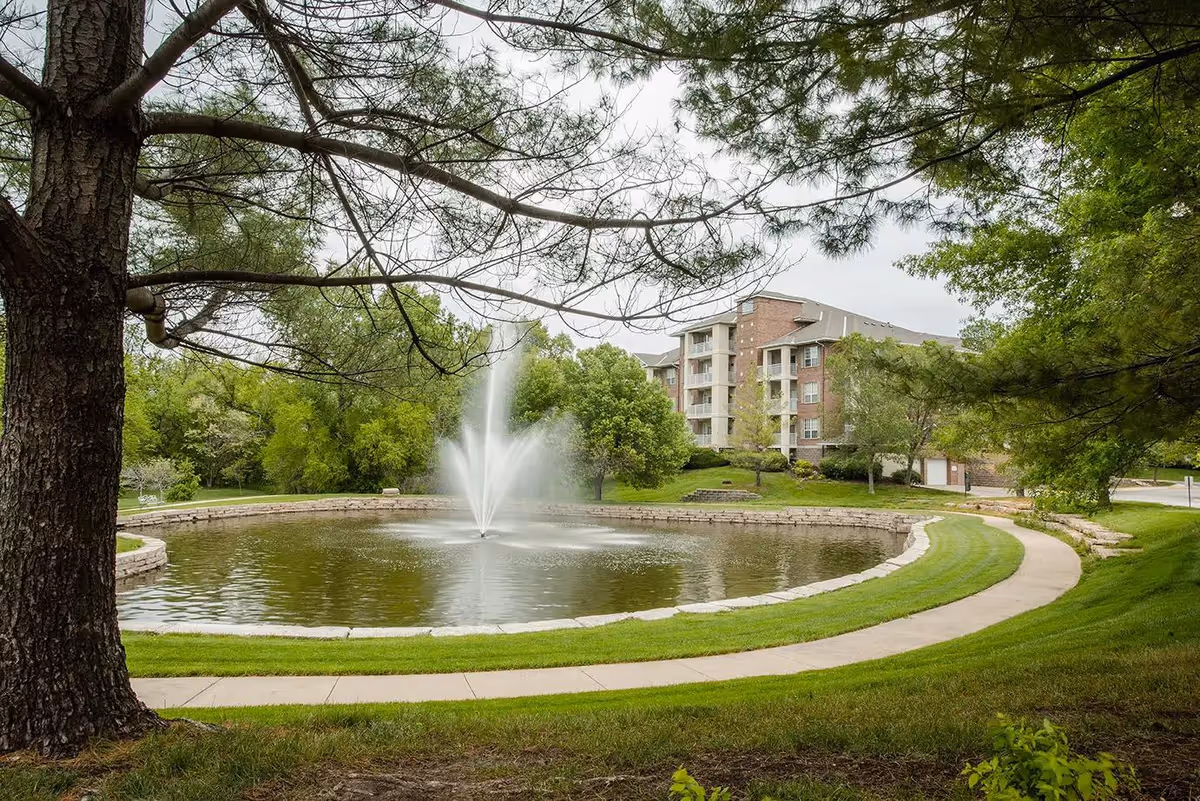 A serene outdoor scene at Delmar Gardens of Overland Park featuring a circular pond with a central water fountain, surrounded by a curved walking path and lush green grass. Trees frame the foreground and background, with a multi-story residential building visible behind the pond.