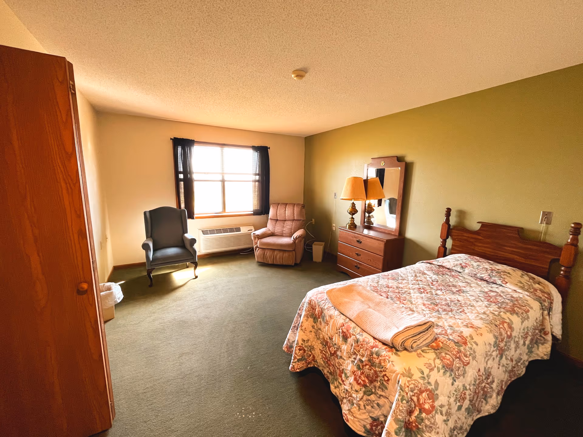 A furnished bedroom with a single bed in floral bedding, a dresser with mirror and lamps, two chairs by a window, and green carpeting.