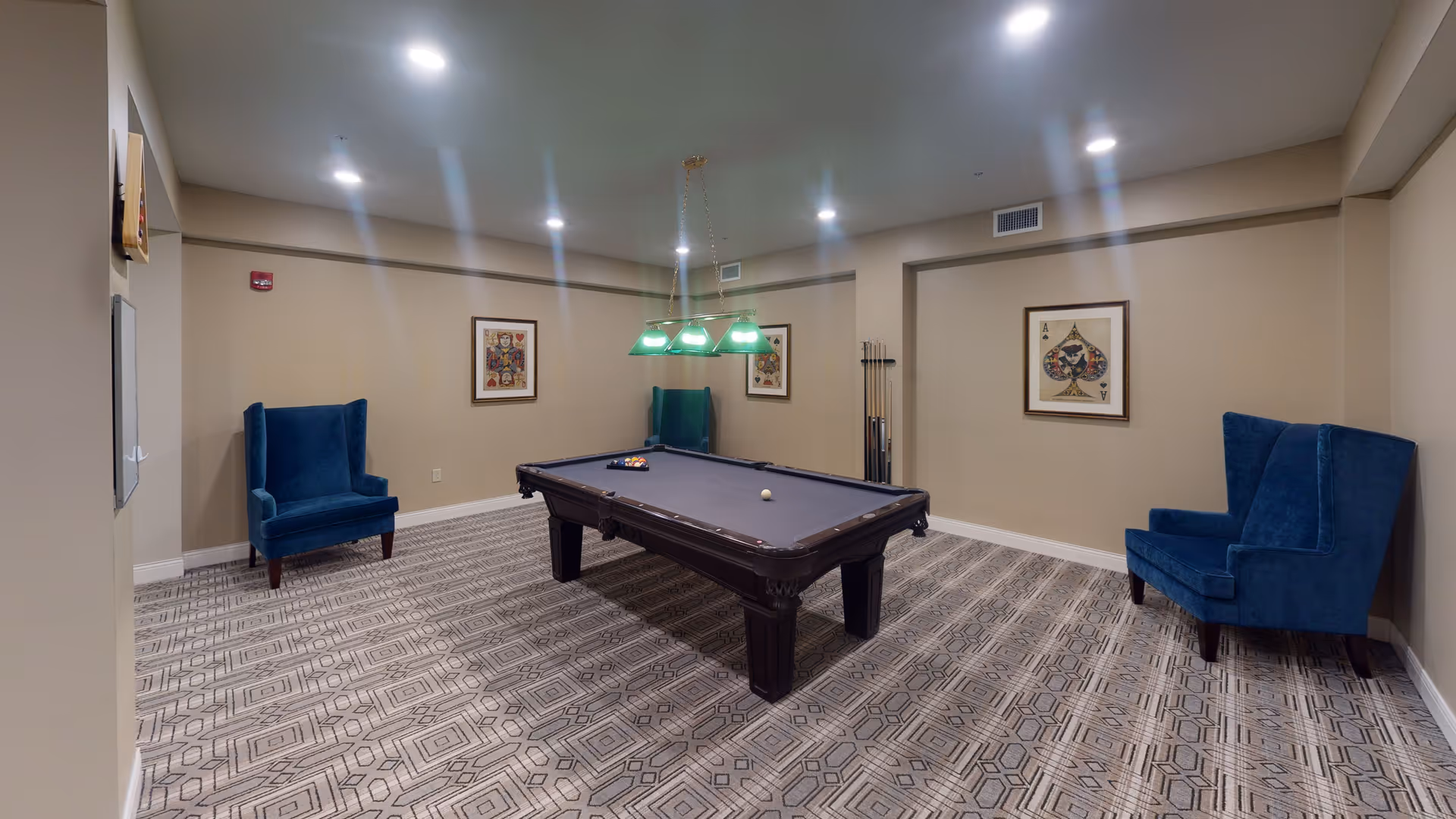 A game room with a pool table in the center, two blue upholstered armchairs placed against the beige walls, three framed playing card artworks on the walls, a patterned carpeted floor, and a green hanging light fixture above the pool table.