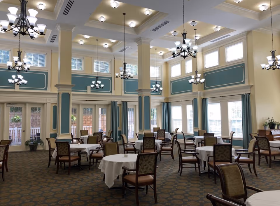 Bright, airy dining room with round tables draped in white cloths, wooden chairs, tall columns, and multiple chandeliers.