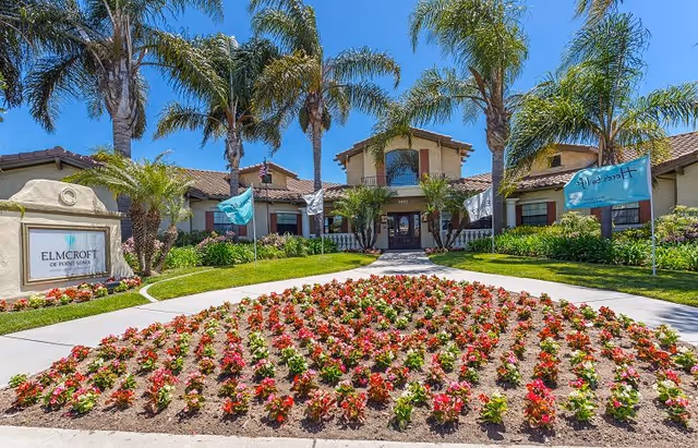 Front exterior view of a senior living facility named Elmcroft of Point Loma with a well-maintained garden featuring rows of colorful flowers, palm trees, and a clear blue sky.