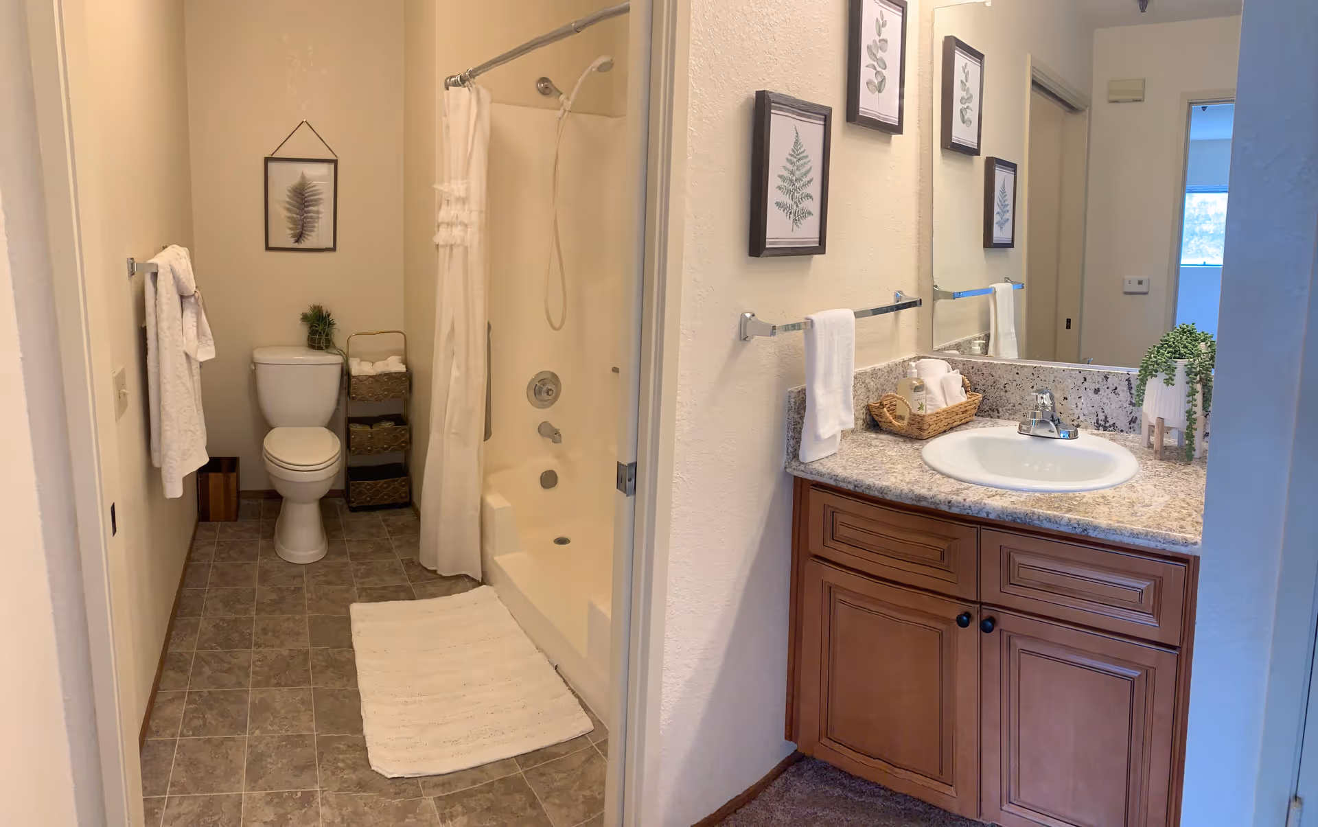 A clean and well-lit bathroom featuring a toilet, a shower with a white curtain, a white bath mat on the tiled floor, and a wooden vanity with a granite countertop and a sink. The walls are decorated with framed botanical prints, and there are towels hanging on a rack and a small basket with toiletries on the counter.