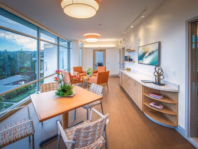 Bright communal dining and lounge area with a wooden table, patterned chairs, orange armchairs, large windows, and a built-in sideboard.