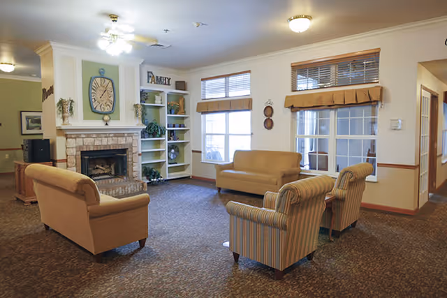 A cozy living room area with a brick fireplace, built-in shelves decorated with plants and ornaments, a large clock above the fireplace, and a sign that says 'FAMILY'. The room has two large windows with blinds and valances, two beige sofas, and two striped armchairs arranged around the fireplace on a carpeted floor.