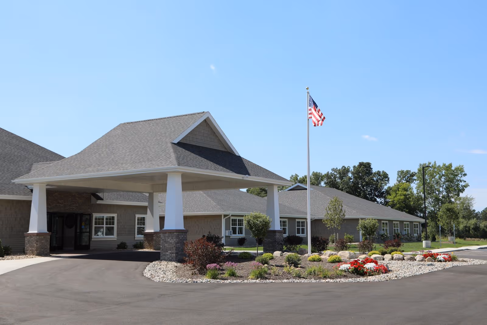 Exterior view of The Brook of Portland facility showing a single-story building with a covered entrance supported by white columns. There is a landscaped area with flowers and shrubs in front, and an American flag on a flagpole. The sky is clear and blue.