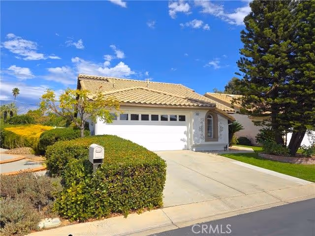 Single-story stucco house with a tiled roof, two-car garage, driveway and manicured shrubs under a blue sky.