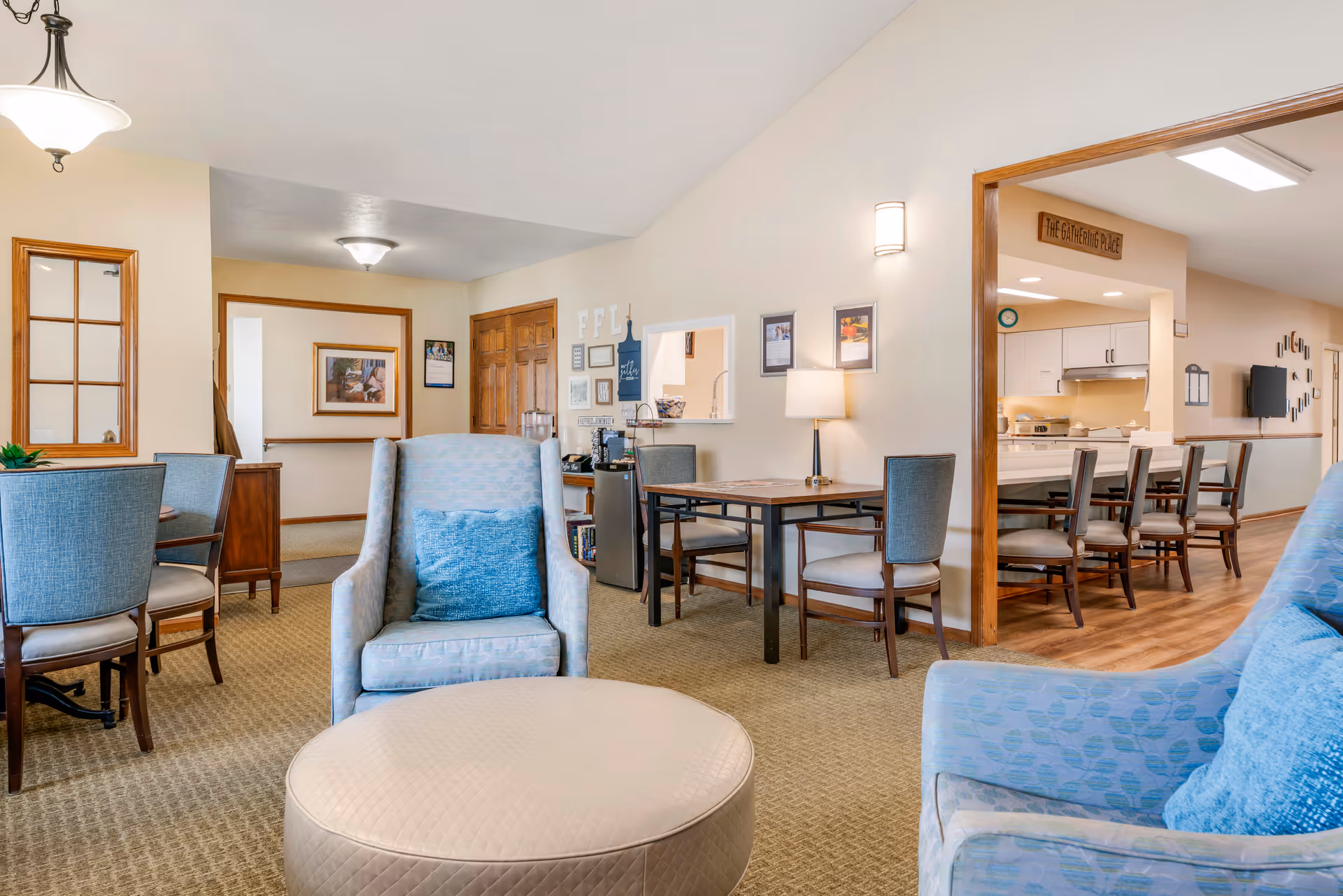 Interior view of a senior living facility lounge area with light blue upholstered armchairs, a round beige ottoman, and several dining tables with chairs. The room has beige walls and carpet, with a kitchen area visible in the background featuring a counter with bar stools. Wall decorations and a lamp on a table add to the cozy atmosphere.