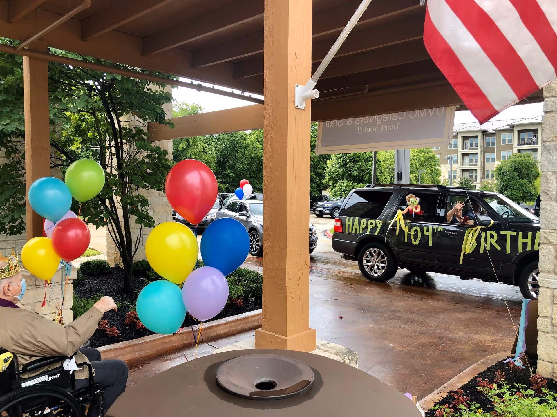 An elderly person in a wheelchair wearing a crown and a face mask is holding colorful balloons under a covered outdoor area. A black SUV decorated with yellow letters spelling 'HAPPY 104TH BIRTHDAY' is parked nearby, with a person inside waving. An American flag is hanging from the wooden structure above.