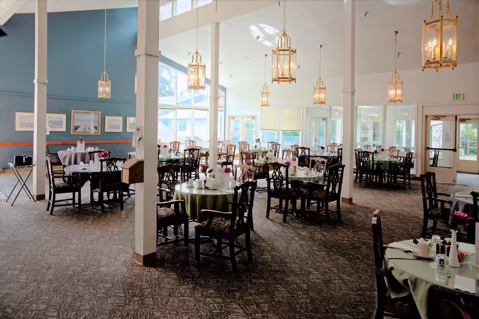 A spacious dining room with multiple round tables covered with green and gray tablecloths, each set with cups, plates, and small flower arrangements. The room features large windows allowing natural light to fill the space, elegant hanging lantern-style light fixtures, and framed artwork on the blue walls. The carpeted floor and wooden chairs add a warm and inviting atmosphere.