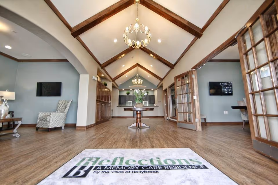 Spacious communal lounge with a vaulted wood-beamed ceiling, chandeliers, seating areas, a central table with flowers, and an entry mat in the foreground.