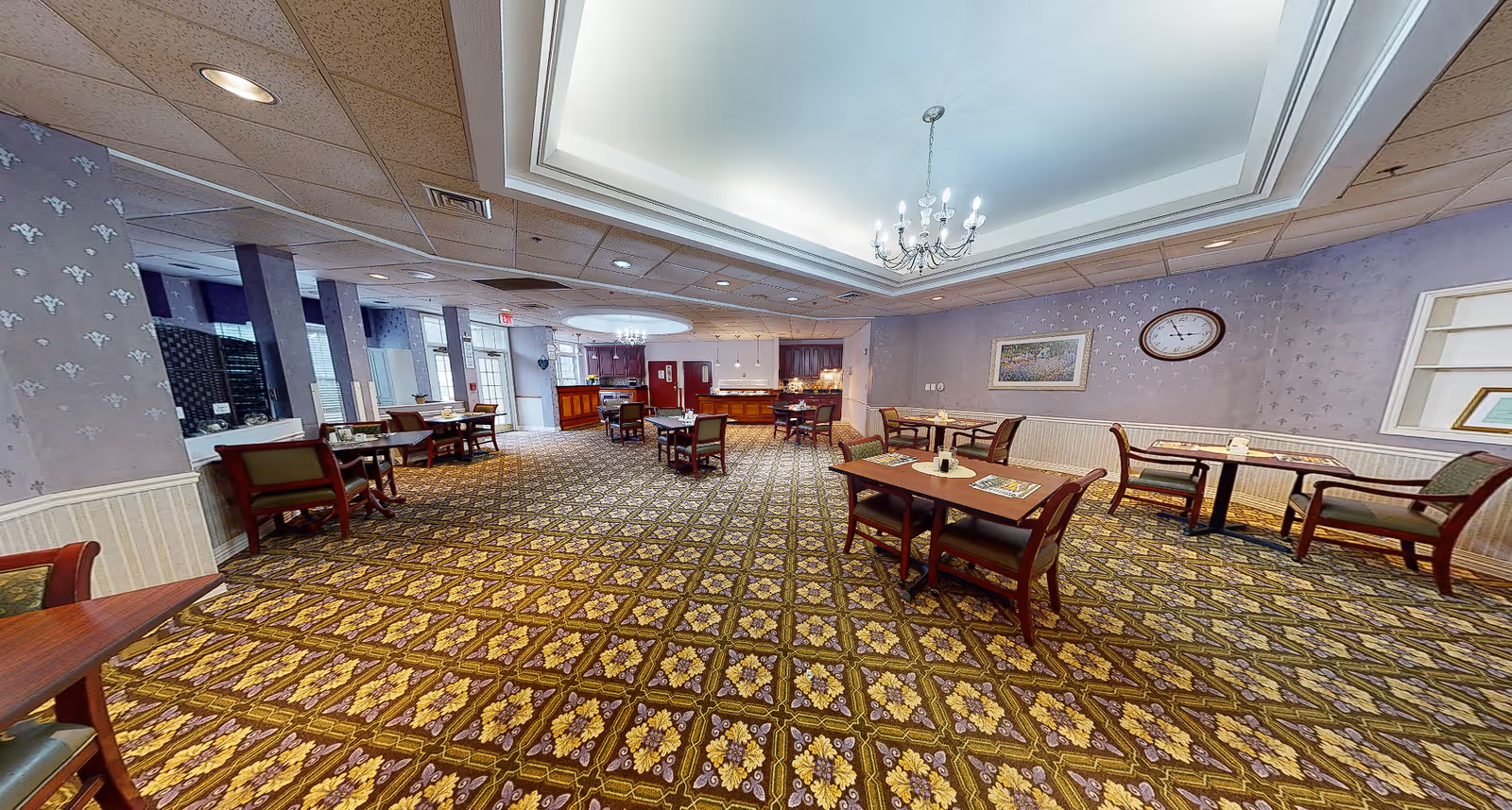 Spacious carpeted dining room with multiple tables and chairs, a chandelier, and a service counter in the background.