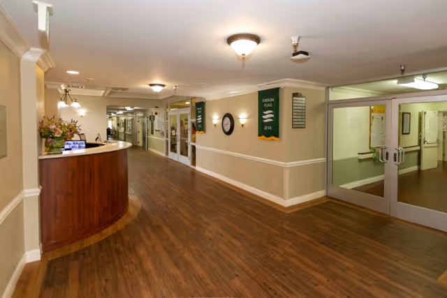 Interior hallway of Camarillo Healthcare Center with wood flooring, beige walls, a curved wooden reception desk with flowers, wall-mounted clock, green banners, and double glass doors leading to another area.