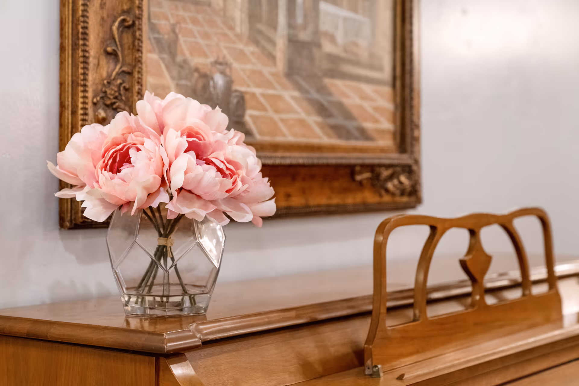 A glass vase of pink peonies sits on a wooden piano beneath a framed painting.