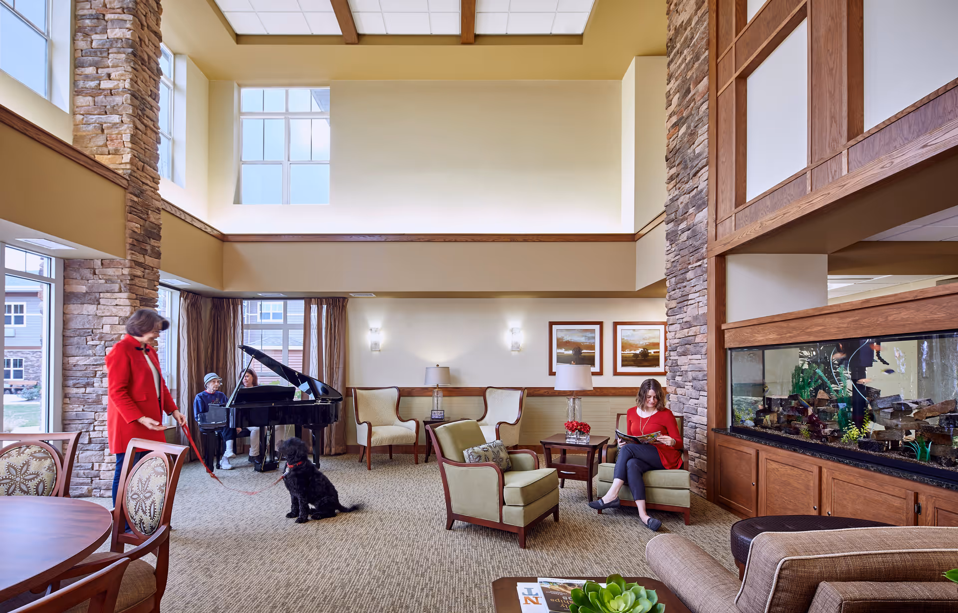 A spacious and well-lit common area in a senior living facility with high ceilings and large windows. The room features a grand piano with two people seated nearby, a woman in a red jacket playing with a black dog, and another woman sitting on a green armchair reading a book. The room is decorated with comfortable chairs, a large fish tank, framed artwork on the walls, and stone pillars.