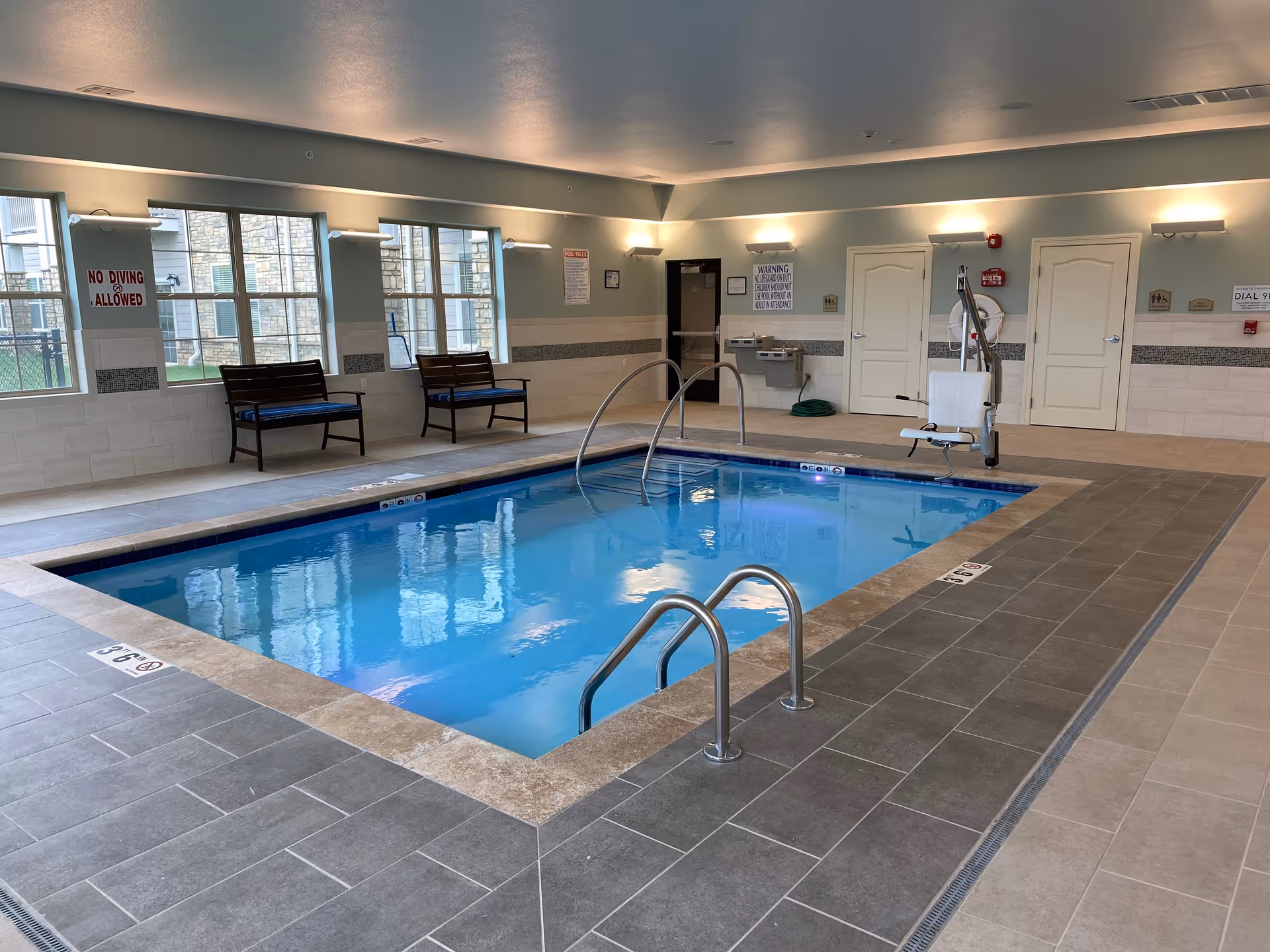 Indoor swimming pool area with steps, handrails, a pool lift and benches next to windows.