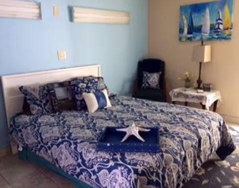 Neatly made bed with blue patterned bedding in a small bedroom with a light blue accent wall, nightstand, lamp and chair.