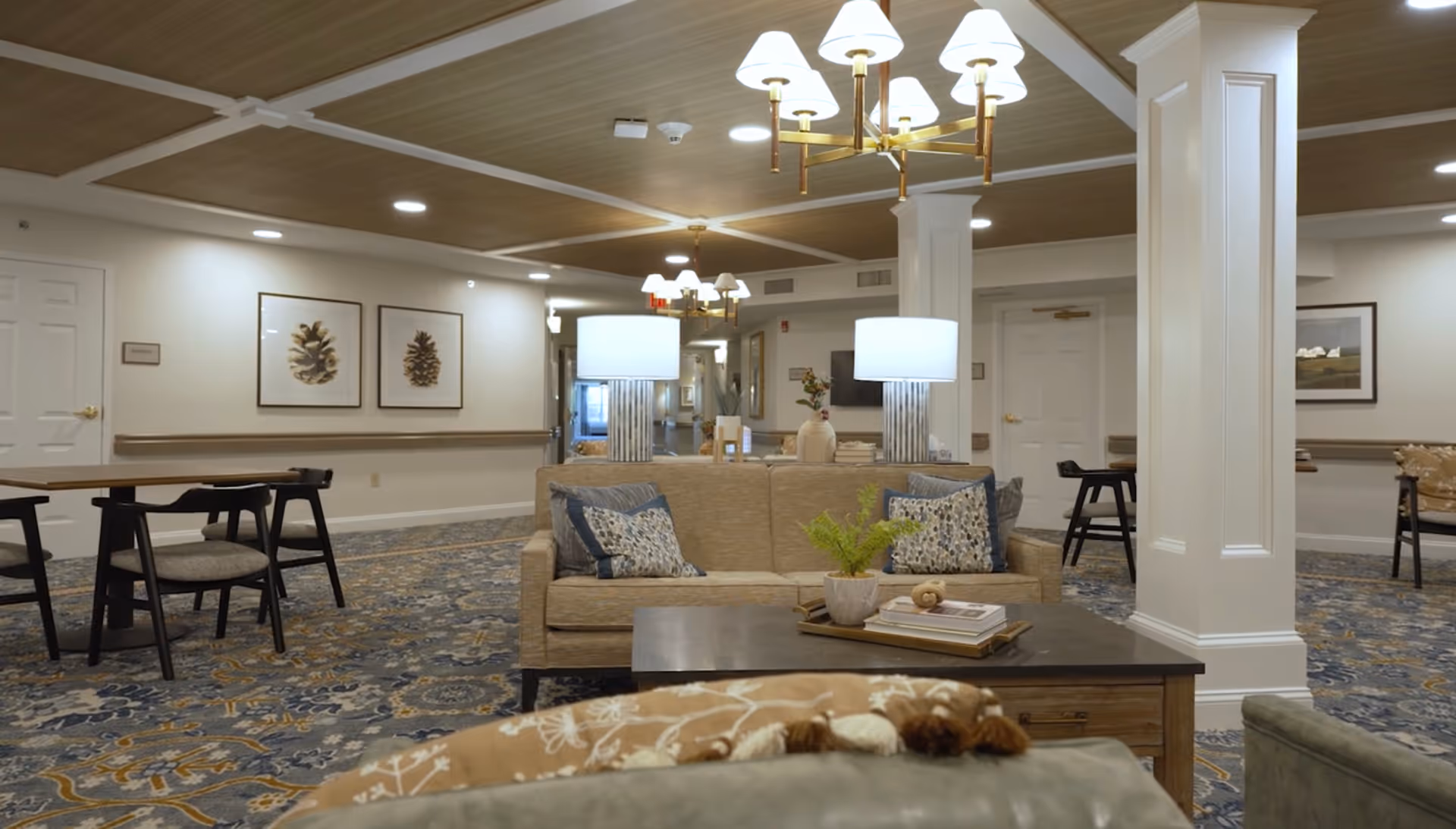 Spacious furnished common lounge with sofas, a coffee table, lamps, chandeliers and dining tables in a senior living facility.