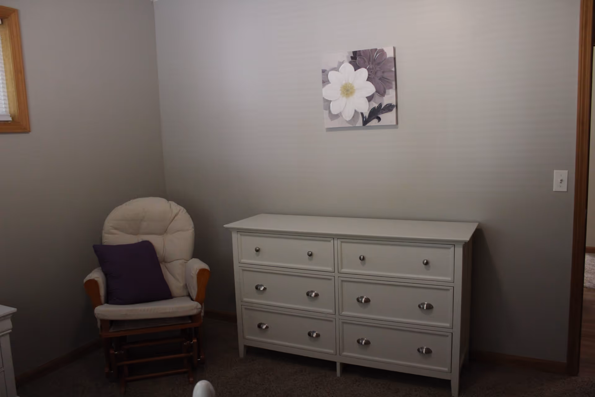 A corner of a bedroom featuring a white cushioned rocking chair with a purple pillow, a white dresser with six drawers, and a floral painting with white and purple flowers hanging on a light gray wall.