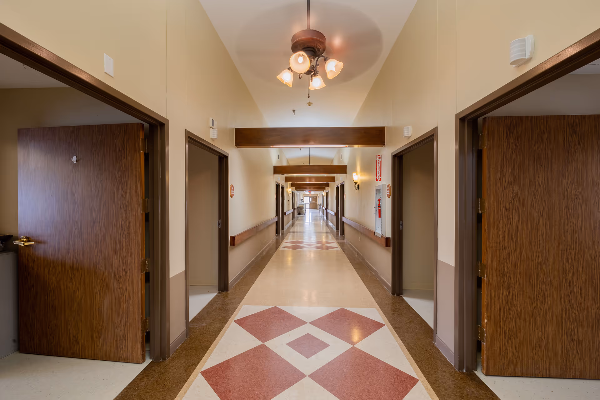 Long, well-lit interior hallway of a nursing and rehabilitation center with open wooden doors, patterned tile floor, ceiling lights and handrails.