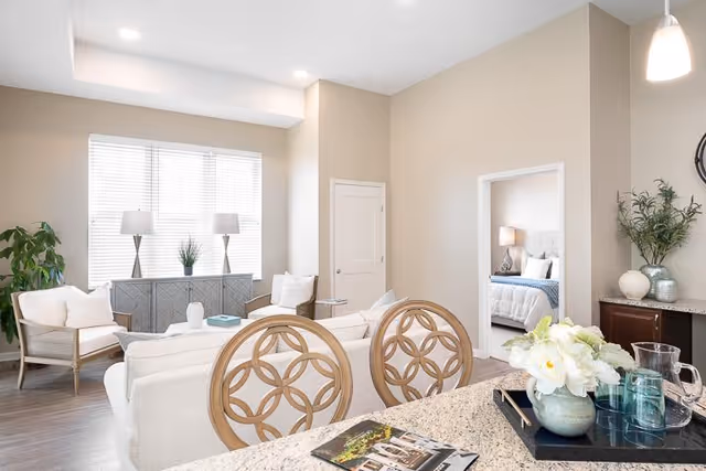 Bright and airy living room area with beige walls and wooden flooring, featuring a white sofa, two armchairs, a sideboard with two lamps and a plant, and a kitchen counter with decorative chairs and a flower arrangement. A bedroom with a bed and nightstand is visible through an open doorway.