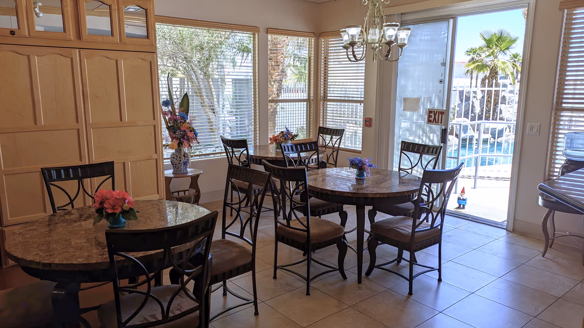 Sunlit dining room with round marble-top tables and chairs, floral centerpieces, and a glass door leading to a pool area.
