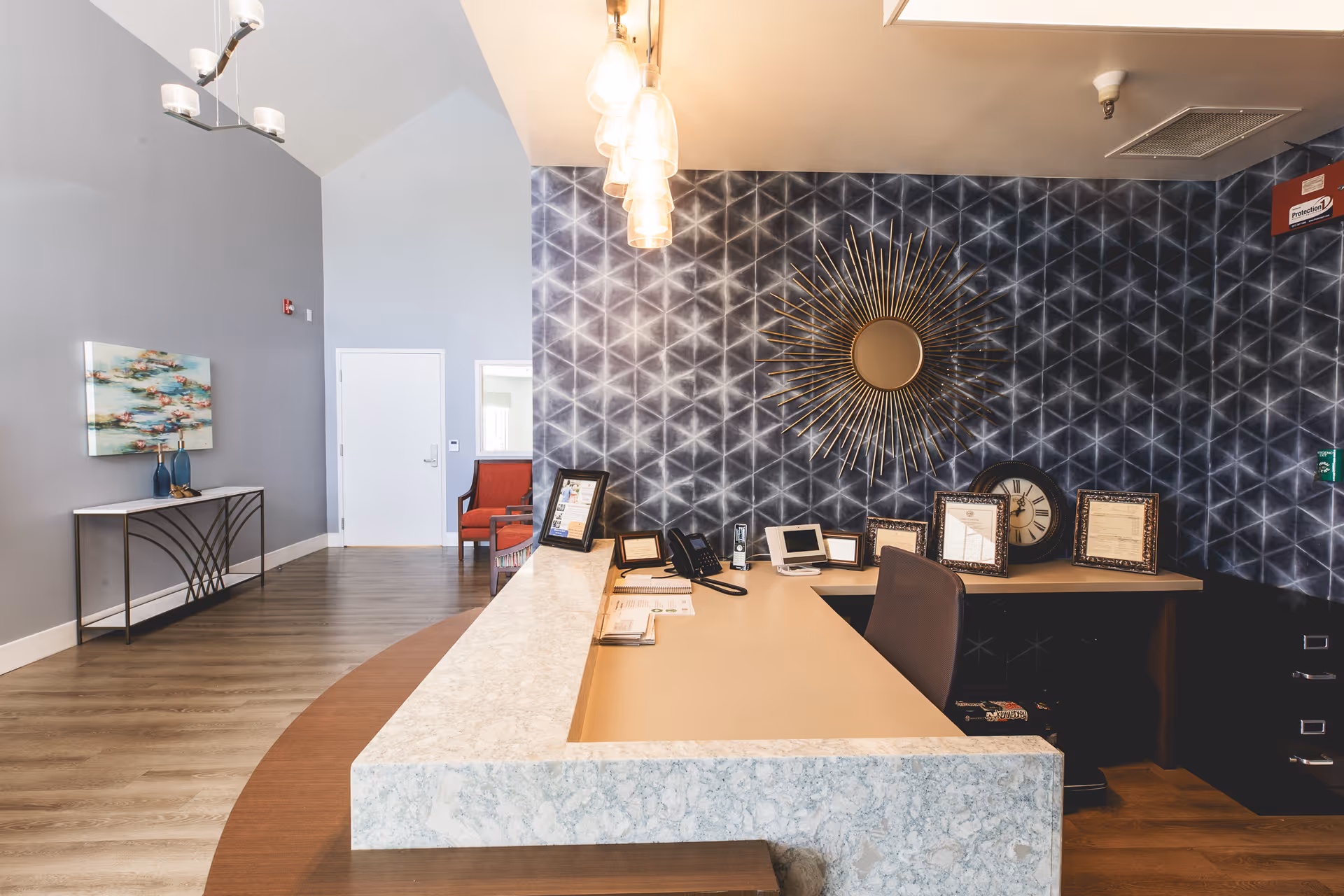 Reception area with a marble countertop desk, a chair, several framed pictures, a telephone, and a decorative sunburst mirror on a patterned dark wall. The room has light wood flooring, a gray wall with a painting, and a red chair near a white door.