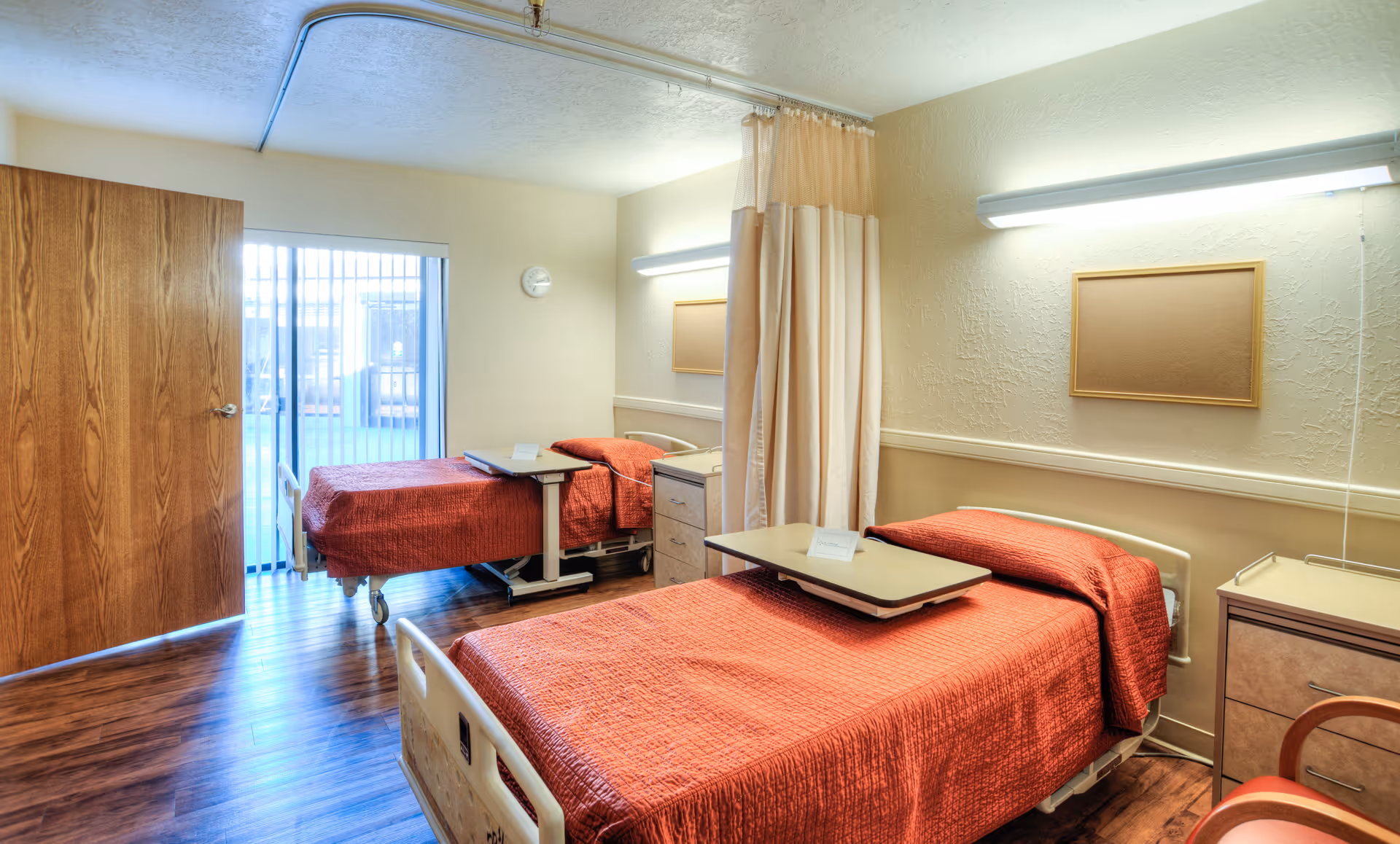 Two hospital-style beds with red bedspreads in a shared patient room with wood flooring and a privacy curtain.