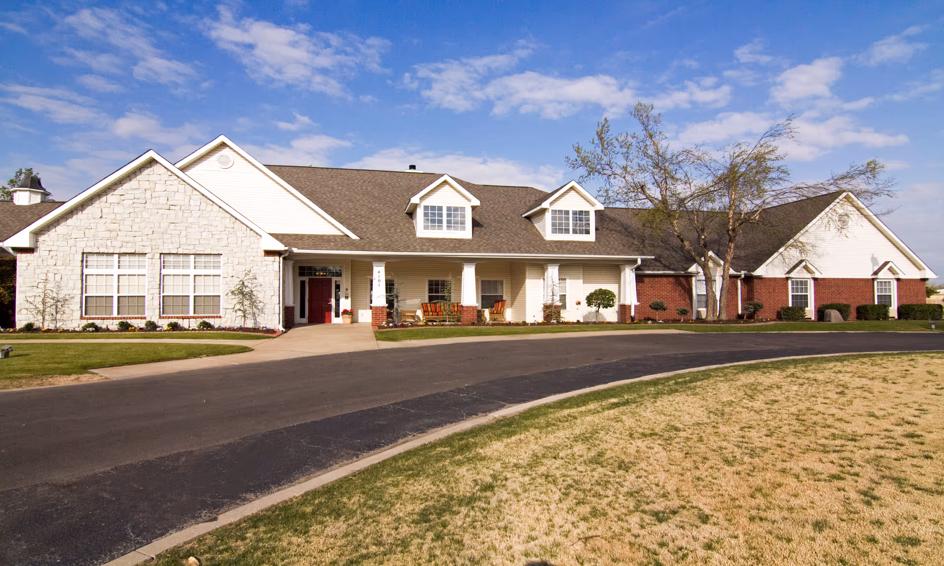 Exterior view of a single-story assisted living facility building with a combination of stone, brick, and siding on the facade. The building has multiple windows, a covered entrance with a red door, and a small porch area with seating. There is a paved driveway and a grassy area in front under a partly cloudy sky.