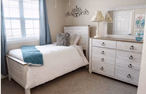 A bright bedroom with a single bed featuring white bedding, a teal throw blanket, and a gray decorative pillow. Next to the bed is a white wooden dresser with a matching table lamp and a large mirror above it. A window with blue curtains allows natural light to fill the room.