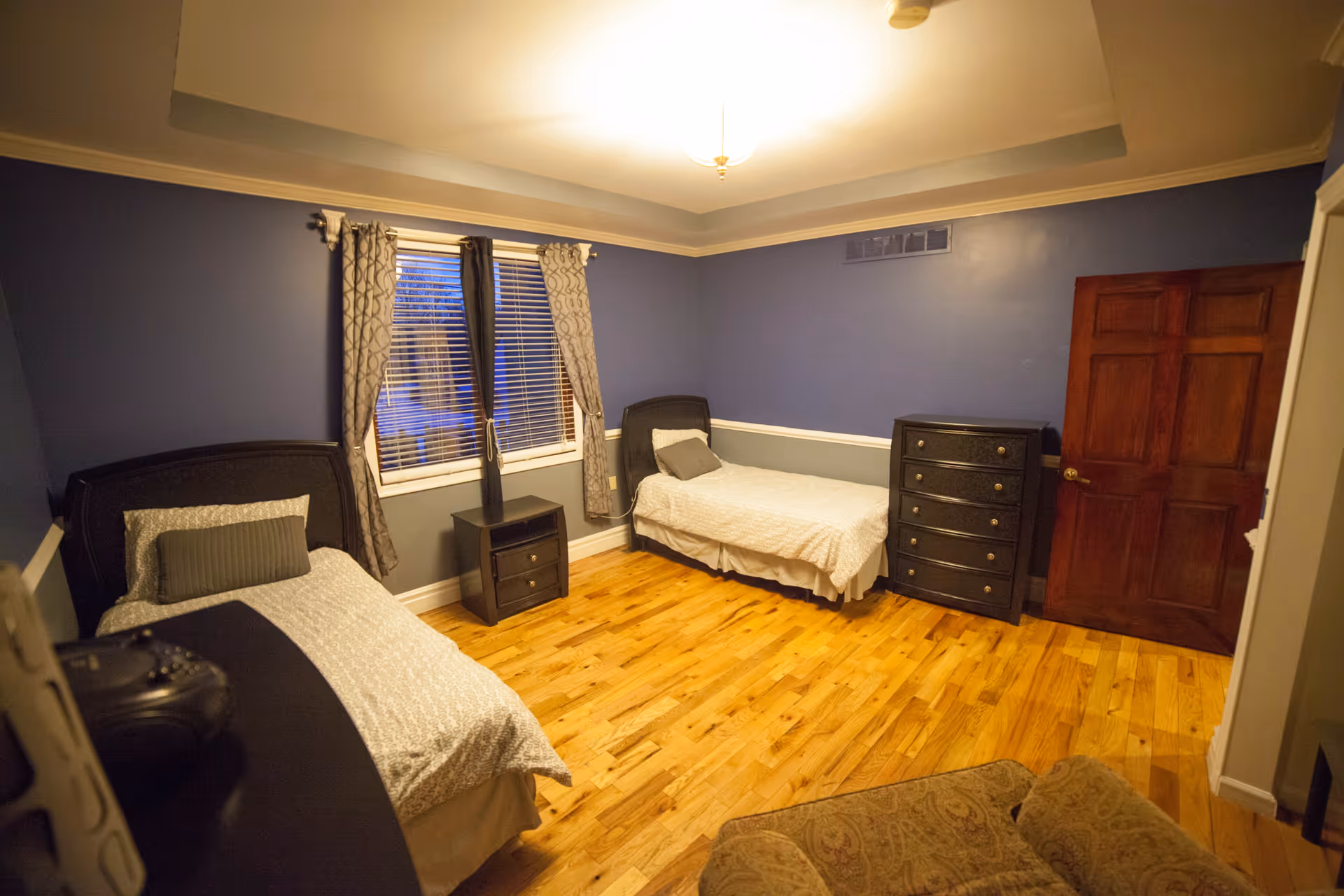A bedroom with two single beds, each with a pillow and light-colored bedding. Between the beds is a small nightstand with a decorative vase. There is a wooden dresser against the wall near a closed wooden door. The walls are painted blue with white trim, and the floor is hardwood. A window with patterned curtains is centered on the wall behind the beds. A cushioned armchair is partially visible in the foreground.