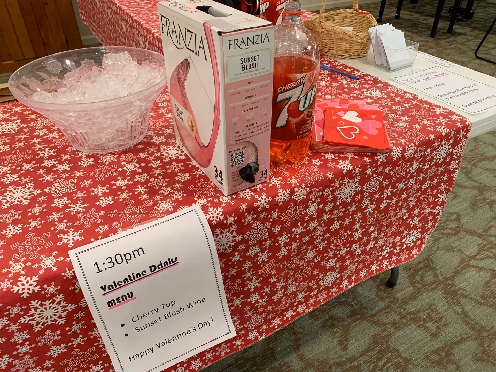 A table with a red snowflake tablecloth holding a box of Franzia wine, a bottle of Cherry 7Up, an ice bowl, and a printed Valentine’s drink menu.