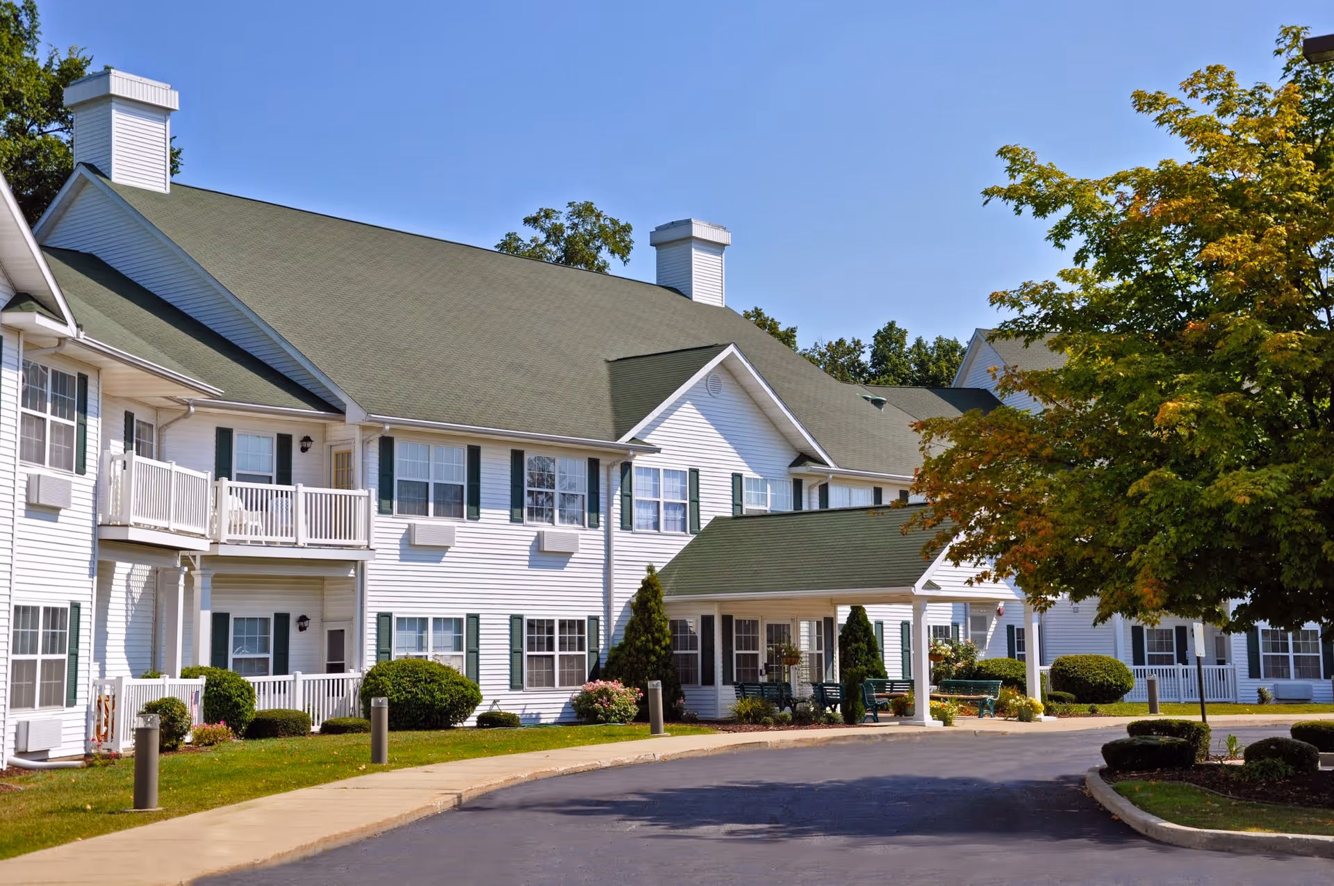 Front exterior of a white two-story senior living building with a green roof, covered entrance, balconies, and landscaped driveway.