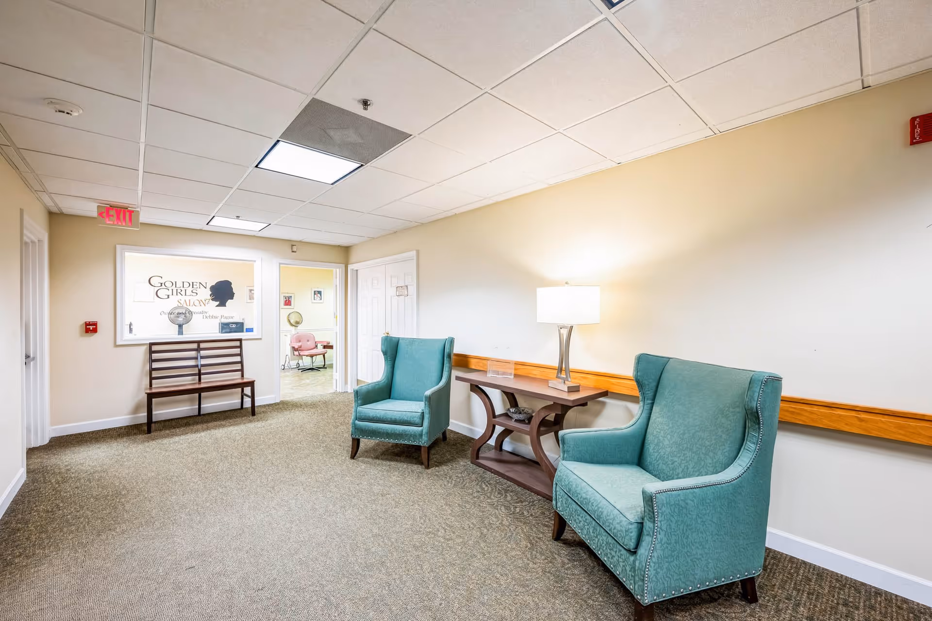 A hallway seating area in a senior living facility with two teal upholstered armchairs, a wooden side table with a lamp, and a wooden bench near a window displaying the sign 'Golden Girls Salon'. The walls are light beige, and the floor is carpeted. There is an exit sign and a fire alarm on the walls.