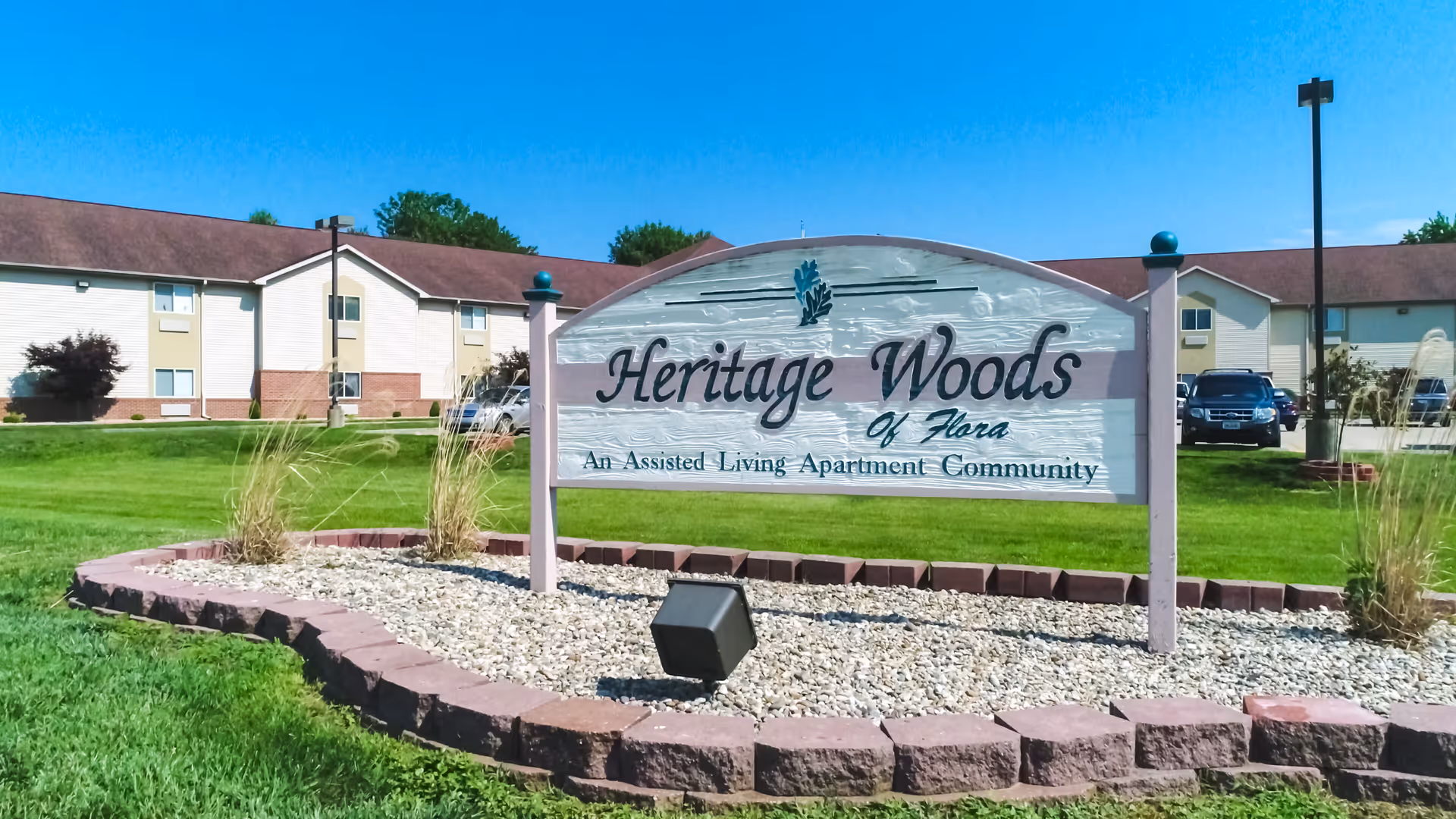 Outdoor view of the Heritage Woods of Flora assisted living apartment community sign with a landscaped area of rocks and plants in front, and the apartment buildings and parked cars visible in the background under a clear blue sky.