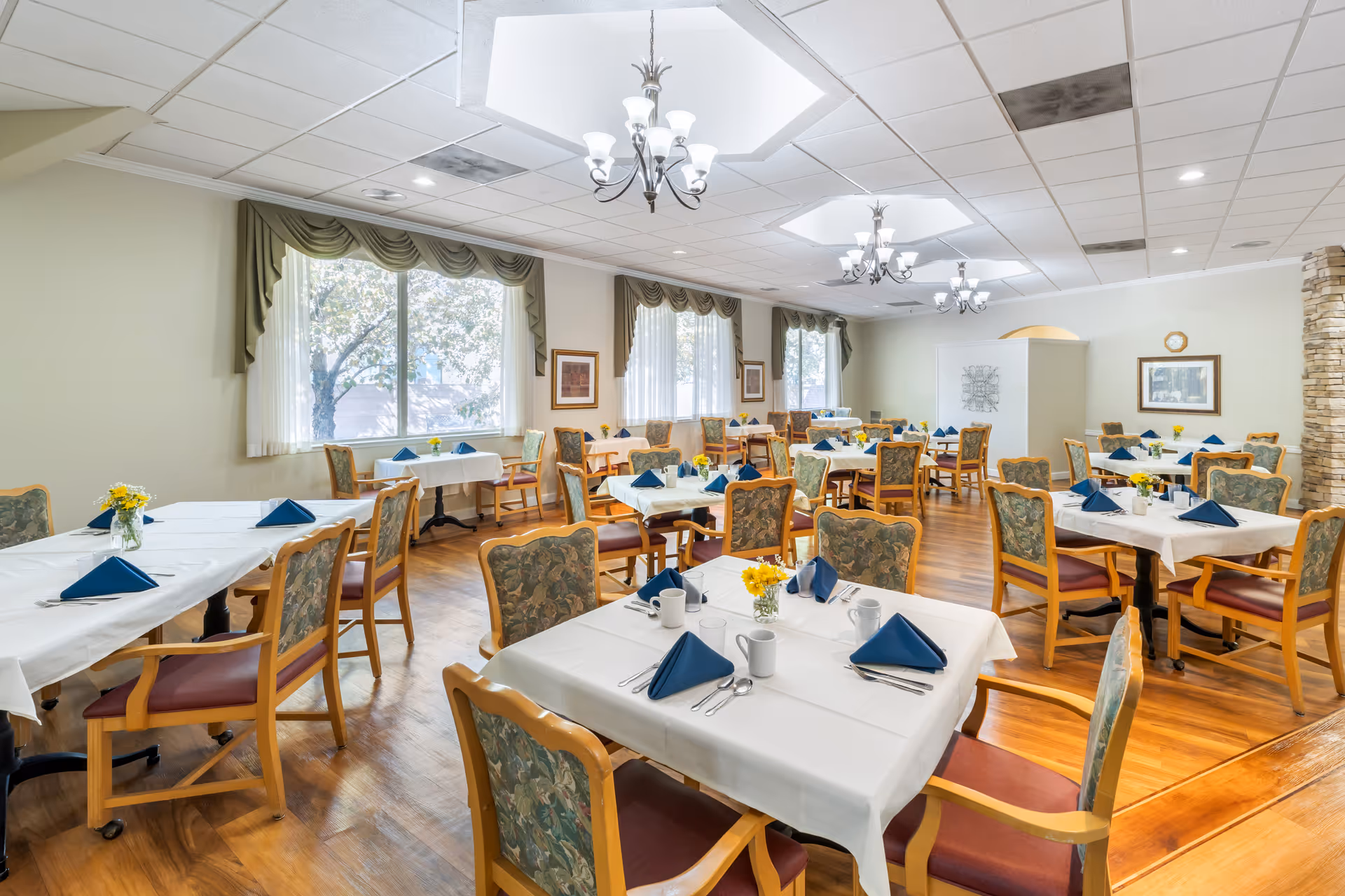 A bright and spacious dining room with multiple tables covered in white tablecloths, each set with blue folded napkins, silverware, mugs, and small vases of yellow flowers. The room has large windows with green drapes, wooden chairs with floral upholstery, and chandeliers hanging from the ceiling.