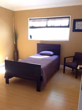 A simple bedroom with a single dark wooden bed covered with a purple bedspread and a matching pillow. Next to the bed is a dark wooden chair with armrests and a small table. The room has light wooden flooring, beige walls, a window with blinds, and a tall decorative vase with long sticks in the corner.