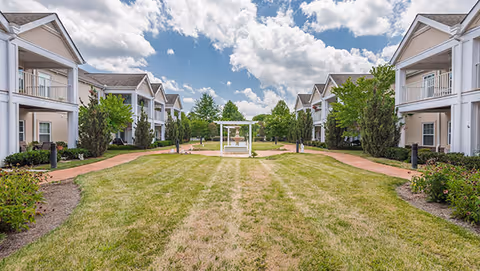 A spacious outdoor courtyard area at Park Place of West Knoxville featuring a well-maintained grassy lawn, a central white pergola with a fountain underneath, surrounded by two-story residential buildings with balconies on either side, under a partly cloudy blue sky.