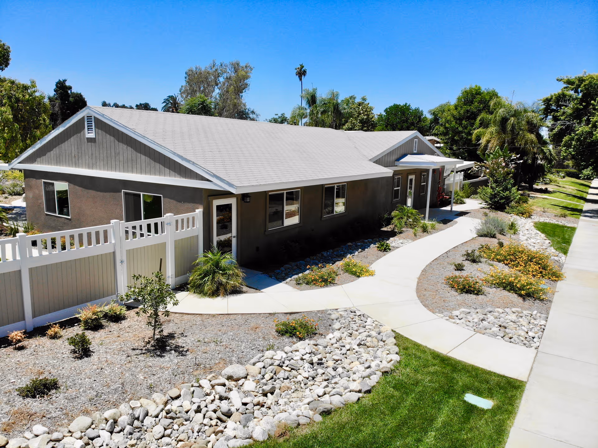 Exterior view of a single-story building with a gray roof and brown walls, surrounded by landscaped gardens with rocks, small plants, and a curved concrete walkway leading to the entrance. The sky is clear and blue, and there are trees and greenery in the background.