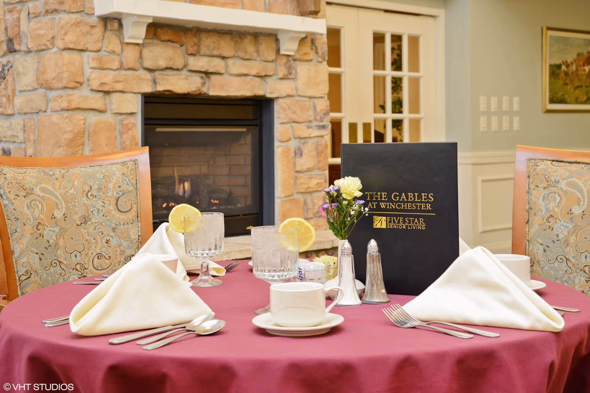A dining table set with a burgundy tablecloth, white folded napkins, silverware, two glasses of water with lemon slices, a coffee cup, salt and pepper shakers, a small flower arrangement, and a black menu folder labeled 'The Gables at Winchester Five Star Senior Living' in front of a stone fireplace and upholstered chairs.