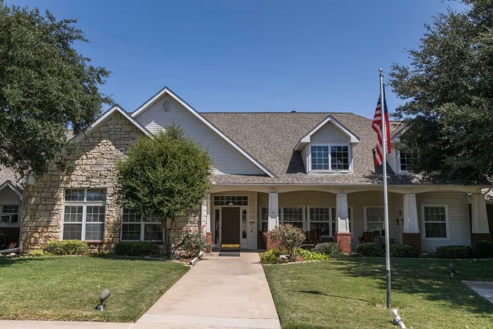 Front exterior view of a senior living facility building with a stone and siding facade, a covered porch with columns, an American flag on a flagpole, and well-maintained green lawn and trees under a clear blue sky.