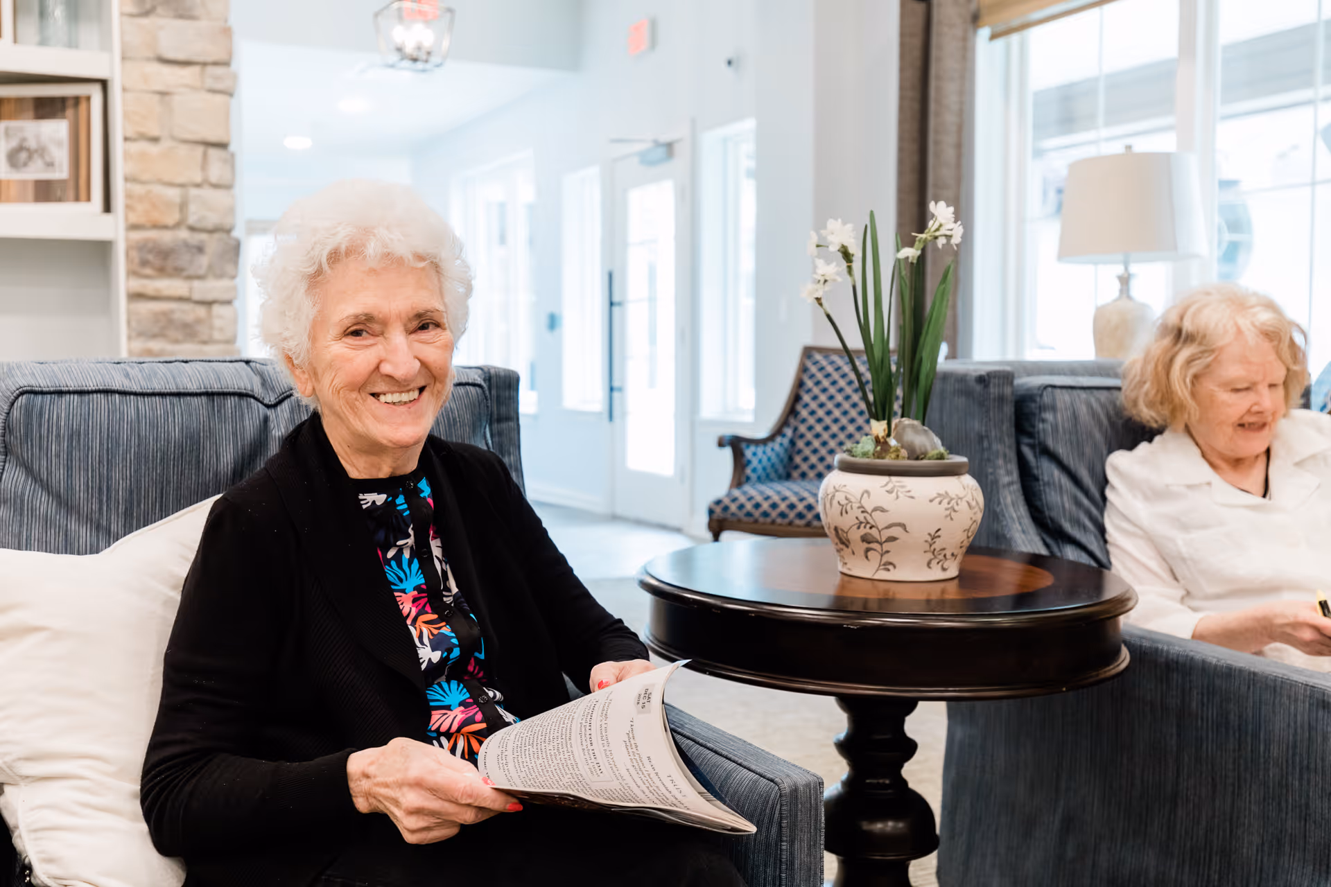 Two elderly women sitting in a bright, comfortable living room area. One woman with white hair is smiling at the camera while holding a newspaper, and the other woman with blonde hair is sitting on a couch in the background. The room features blue upholstered chairs, a round wooden table with a decorative flower pot, large windows, and a lamp.