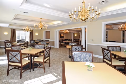 A spacious dining room with multiple square tables and cushioned chairs arranged neatly. The room features beige walls, carpeted floors, large windows letting in natural light, and elegant gold chandeliers hanging from the ceiling. There are decorative columns and a large mirror on one wall.