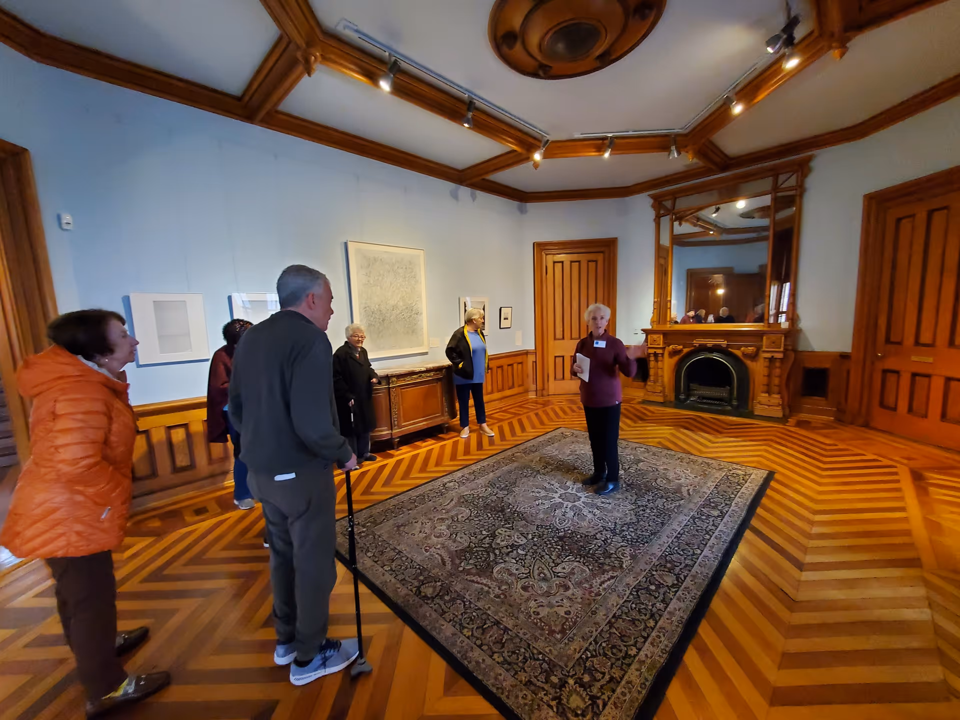 A group of elderly people standing in a spacious room with wooden floors and wood-paneled walls. One woman appears to be speaking or presenting to the group. The room features a large ornate mirror above a fireplace, framed artwork on the walls, and a patterned area rug in the center.