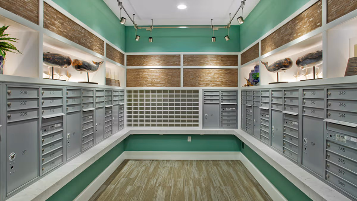 Mailroom interior with rows of numbered silver mailboxes lining three walls beneath teal-painted walls and decorative shelving.